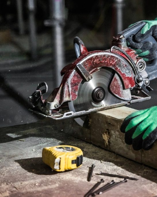 Behandschuhte Hände führen eine Handkreissäge durch einen Holzbalken, Späne fliegen